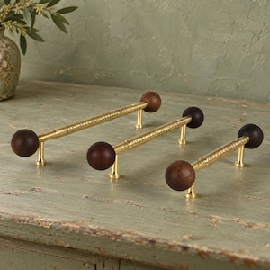 May include: Three cabinet pulls featuring a horizontal brass bar with a dark brown wooden sphere at each end. Each pull is supported by two small brass posts. The text Abarrotesjuanki is visible across the center of each brass bar.