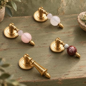 May include: Four brass drawer pulls with circular backplates. Three pulls feature spherical stone accents in pale pink, translucent white, and mottled dark red. One pull is solid brass with a tapered handle. Each pull has a pointed finial tip.
