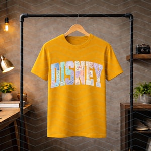 May include: A mustard yellow short-sleeve t-shirt featuring the word DISNEY printed across the chest in a large, arched, block-letter font. Each letter is filled with a unique, colorful patchwork pattern of floral, plaid, and geometric designs.
