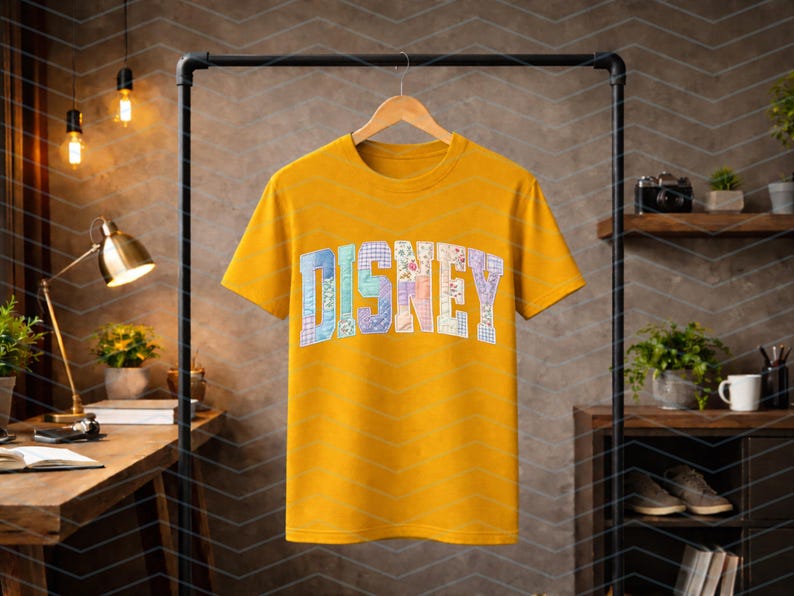 May include: A mustard yellow short-sleeve t-shirt featuring the word DISNEY printed across the chest in a large, arched, block-letter font. Each letter is filled with a unique, colorful patchwork pattern of floral, plaid, and geometric designs.