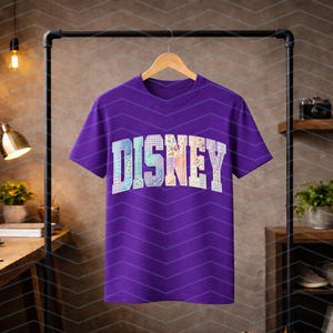 May include: A short-sleeved purple t-shirt featuring the word DISNEY printed across the chest in a large, arched, collegiate-style font. Each letter is filled with a unique, multi-colored floral and plaid pattern in pastel shades.