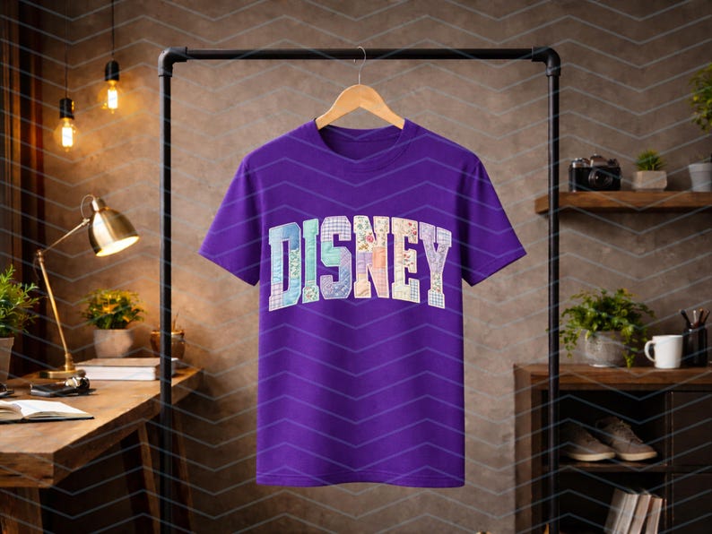 May include: A short-sleeved purple t-shirt featuring the word DISNEY printed across the chest in a large, arched, collegiate-style font. Each letter is filled with a unique, multi-colored floral and plaid pattern in pastel shades.