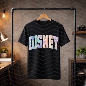 May include: A black short-sleeve t-shirt featuring the word DISNEY in large, block-style letters across the chest. Each letter is filled with a unique, colorful floral and plaid patchwork pattern in pastel shades of blue, pink, and yellow.