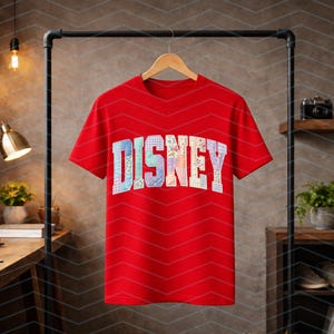 May include: A bright red short-sleeved t-shirt featuring the word DISNEY printed across the chest in large, block-style letters. Each letter is filled with a unique, colorful patchwork pattern consisting of various floral and geometric designs.