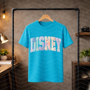 May include: A bright turquoise short-sleeve t-shirt featuring the word DISNEY printed across the chest in a large, arched block font. Each letter is filled with a unique, colorful floral and plaid patchwork pattern in various pastel shades.