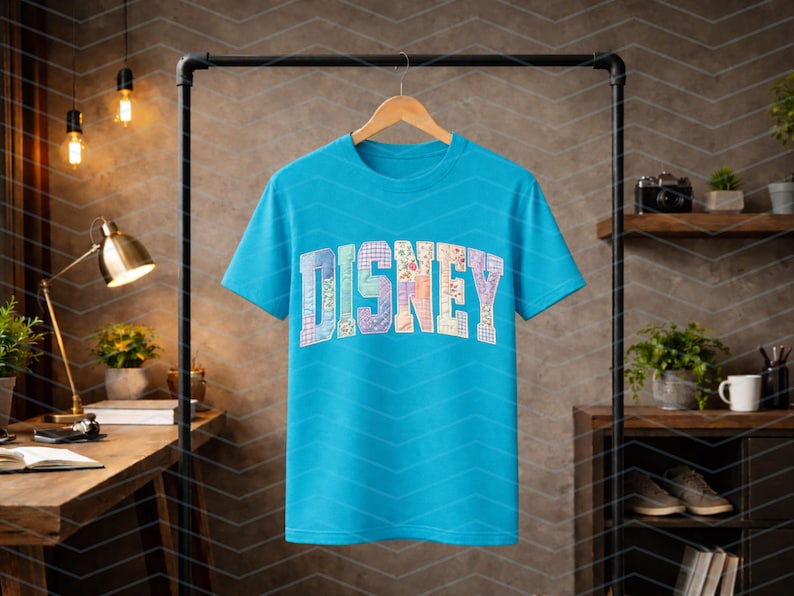 May include: A bright turquoise short-sleeve t-shirt featuring the word DISNEY printed across the chest in a large, arched block font. Each letter is filled with a unique, colorful floral and plaid patchwork pattern in various pastel shades.