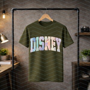 May include: An olive green short-sleeve t-shirt featuring the word DISNEY printed across the chest in a block font. Each letter is filled with a different pastel-colored floral and plaid patchwork pattern, creating a multi-colored graphic design.