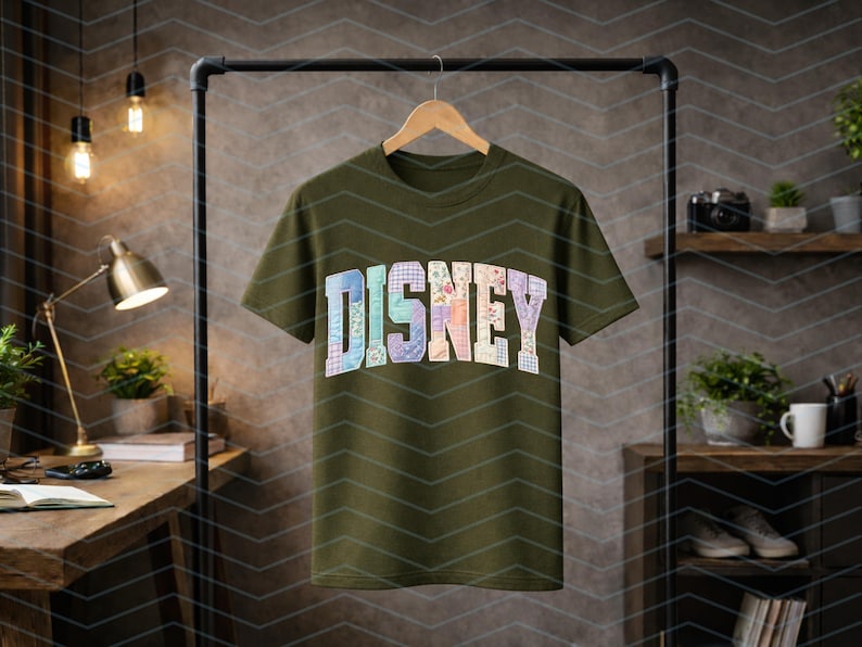 May include: An olive green short-sleeve t-shirt featuring the word DISNEY printed across the chest in a block font. Each letter is filled with a different pastel-colored floral and plaid patchwork pattern, creating a multi-colored graphic design.
