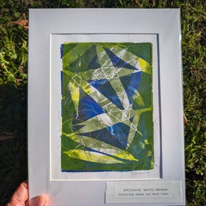 May include: An abstract print featuring layered geometric shapes in shades of green, blue, and white, set within a white mat board. A small label at the bottom reads: Unfiltered, mostly because filtering takes too much time. The artwork is signed.