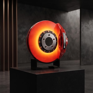 May include: A circular brake disc display piece featuring a glowing orange light effect, a central silver hub with black bolts, and a red brake caliper with the word PORSCHE printed in white lettering, all mounted on a black stand.