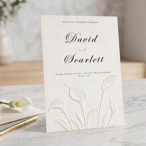 May include: A white wedding invitation card featuring embossed calla lily floral designs at the bottom. The text reads: Join us for a beautiful celebration, David and Scarlett, Saturday, September 12, 2026 at 5:00 PM at The Grand Ballroom, Bloom Hill.