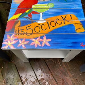 May include: A wooden table painted with a tropical scene. A red parrot sits on a branch with a margarita glass in its beak. The table has a sign that says "it's 5 o'clock!" and is decorated with pink flowers.