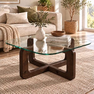 Peut inclure: Une table basse carrée avec un plateau en verre transparent et une base en bois brun foncé au design en croix. La table supporte un vase en céramique claire avec de la verdure, un petit bol en bois et deux livres empilés intitulés The Kinfolk Home et At Home.
