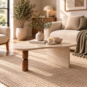 Puede incluir: Una mesa de centro con un tablero de travertino beige de forma orgánica irregular, apoyado sobre una pata cilíndrica de madera oscura y una base de losa de travertino rectangular. La superficie sostiene un libro, una vela y un cuenco.