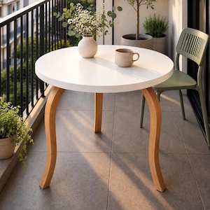 May include: A round white table with three curved light wood legs. On the table sits a white ribbed vase containing white flowers and a beige ceramic mug. Beside the table is a sage green chair with a slatted back and a potted green plant.