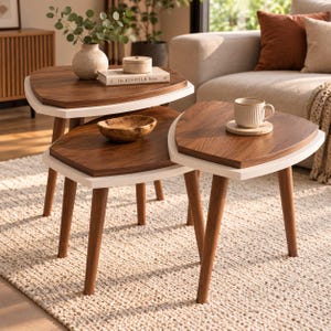 May include: A set of three nesting tables featuring dark brown wooden tops with white edges and tapered wooden legs. The tables are arranged in a tiered formation, with a ceramic vase, a small box, a book titled The Kinfolk Home, and a bowl.