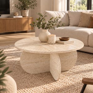 May include: A round coffee table crafted from beige travertine stone with a smooth surface and a cross-shaped base. The table holds a light-colored ceramic vase with green foliage, a textured cylindrical candle, and a small wooden bowl on a book.