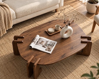 May include: A low-profile, dark brown wooden coffee table with an irregular, rounded shape and four distinct, interlocking leg supports. The tabletop holds an open magazine, a small rectangular wooden tray, a white ceramic mug, and a white vase.