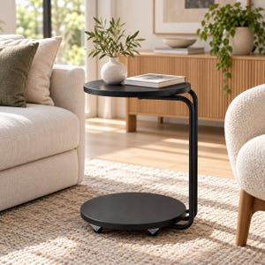May include: A black C-shaped side table with a round top and a round base, featuring a metal frame and small wheels for mobility. The table holds a white speckled vase with green leaves and a closed book on its top surface.