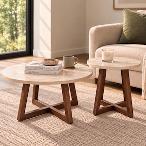 May include: Two round tables with light-coloured stone tops and dark brown wooden legs arranged in a cross-base design. The larger table holds two stacked books titled The Kinfolk Home and At Home, a small bowl, and a ribbed mug on a coaster.