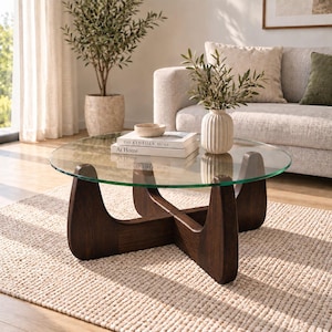 May include: A round coffee table with a clear glass top and a dark brown wooden base featuring a cross-shaped design. The table holds two stacked books titled The Kinfolk Home, a small beige bowl, and a ribbed white vase with green foliage.