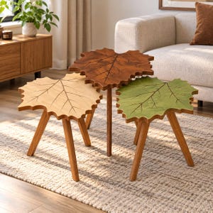 Peut inclure: Un ensemble de trois tables d'appoint en bois en forme de grandes feuilles. Chaque table présente une couleur distincte : bois naturel clair, brun moyen et vert sauge. Chaque table possède une gravure de nervure centrale et trois pieds en bois inclinés.