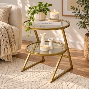 May include: A two-tier round side table with a gold metal frame and clear glass shelves. The top shelf holds a potted pothos plant, a Sandalwood candle, and books titled The Kinfolk Home and At Home. The bottom shelf holds two white candle holders.