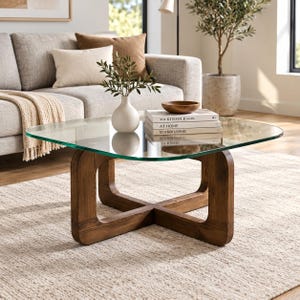 May include: A square coffee table with a clear glass top and a dark brown wooden base featuring a cross-shaped design. The table holds a white ceramic vase with green foliage, a small wooden bowl, and a stack of three books titled The Kinfolk Home, At Home, and Scandi Living.
