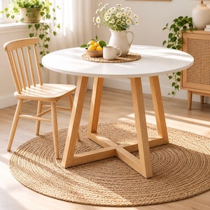 May include: A round white dining table with light wood legs in an X-shaped base, accompanied by a matching light wood spindle-back chair. The table is set with a woven placemat, a white ceramic pitcher of daisies, and a bowl of yellow lemons.