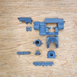 May include: A collection of unpainted grey plastic model kit components, including a large central chassis frame, a cylindrical barrel, a rectangular panel, and several smaller mechanical parts arranged for assembly on a flat surface.