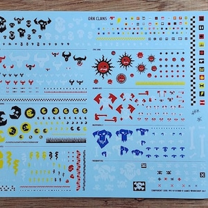 Puede incluir: Una hoja de calcomanías azul claro titulada ORK CLANS que presenta varios iconos gráficos en miniatura, incluyendo calaveras, soles, lunas, hachas, llamas y patrones de cuadros en colores negro, rojo, amarillo, blanco y azul para modelismo.