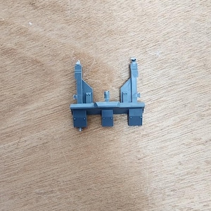 May include: A grey plastic model kit component featuring a horizontal base with three rectangular tabs extending downward and two vertical upright supports rising from the sides, with a small central protrusion between the uprights.