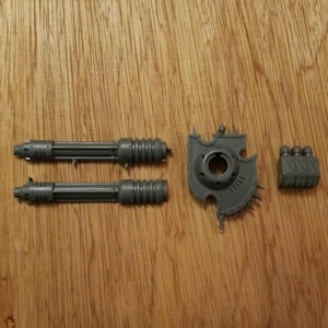Puede incluir: Un conjunto de componentes de modelo de plástico gris sin pintar que incluye dos piezas de cañón cilíndricas largas con detalles acanalados, una placa circular tipo escudo con un orificio central y emblema de calavera, y un bloque rectangular pequeño.