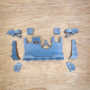 May include: A collection of unpainted grey plastic model kit components, including a large central rectangular panel with a winged skull emblem, two side panels, two mechanical arm pieces, two small square plates, and two circular base connectors.
