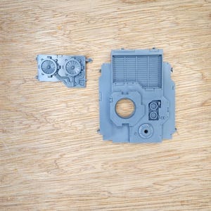 May include: Two unpainted grey plastic model kit components. The smaller piece features two circular fan vents and piping. The larger piece is a rectangular panel with a grid vent, a central circular opening, and small mechanical details.