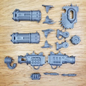 Puede incluir: Una colección de componentes de maquetas de plástico gris sin pintar, que incluye dos cañones cilíndricos estriados, una gran hombrera con púas, varias articulaciones mecánicas, un bloque lanzamisiles y varias piezas de conexión pequeñas.