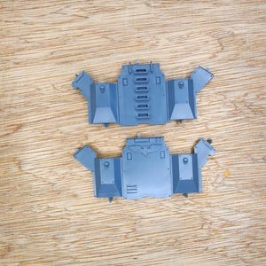 May include: Two grey plastic model kit components featuring angular side panels and central sections. The top piece displays a vertical row of rectangular vents, while the bottom piece features a molded eagle emblem and small skull details on the sides.
