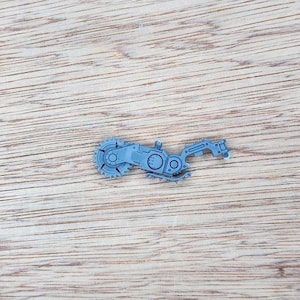 Op de afbeelding: Een klein, grijs plastic miniatuuronderdeel met een cirkelzaagblad aan één uiteinde, verbonden met een mechanische arm met ingewikkelde tandwielachtige details, klinknagels en een gesegmenteerde structuur voor in een modelbouwset.