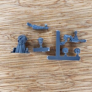 May include: A set of unpainted gray plastic miniature model parts including a torso in a long coat, a separate head with a peaked cap, a detached arm, and a sprue frame containing a weapon arm and two circular shoulder pads with skull emblems.