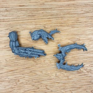 May include: Three unpainted grey plastic miniature components including a muscular arm with a multi-barreled weapon, a small organic-looking shoulder or torso piece, and a curved, serrated blade-like appendage with sharp, pointed protrusions.