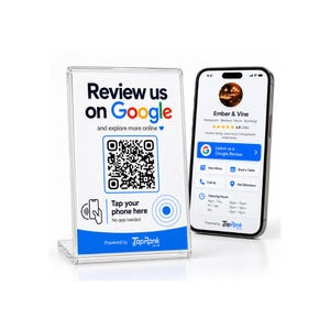 May include: A clear acrylic sign holder displaying a Review us on Google QR code with Tap your phone here instructions, positioned next to a smartphone showing a business profile for Ember & Vine with a Google review button and TapRank branding.