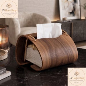 May include: A curved dark wood tissue box holder featuring a smooth grain finish and gold-toned metal handles on the top. The holder contains a stack of white facial tissues that extend through the central opening, showcasing a sleek design.