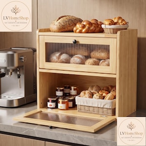 May include: A two-tier wooden bread box with a light natural finish, featuring a top compartment with a fluted glass drop-down door and an open bottom shelf. The unit holds various loaves of bread, a small woven basket, and glass jars.