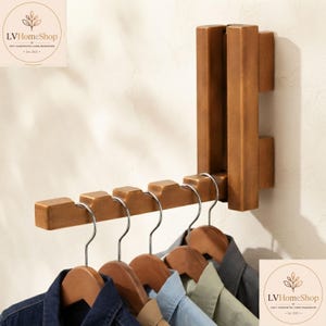 May include: A wall-mounted wooden garment rack with a folding arm featuring five notches for hangers. The rack is finished in a medium brown tone and holds four wooden hangers with shirts in navy, light blue, light green, and dark gray.