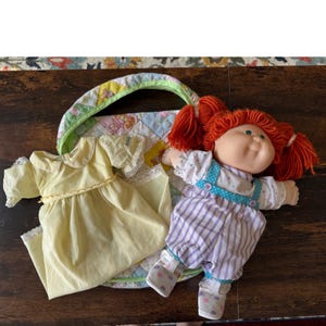 Könnte beinhalten: Eine Puppe mit leuchtend orangefarbenem Wollhaar in Zöpfen, bekleidet mit einem weißen Hemd mit blauen Punkten und einer lila gestreiften Latzhose. Daneben liegen ein hellgelbes Kleid mit Spitzenbesatz und eine gesteppte Tasche mit Pastellmuster.