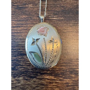 May include: An oval-shaped gold-tone metal locket pendant suspended from a matching rope chain. The front surface features an engraved floral design with a pink-tinted flower at the top and green-tinted leaves at the base, framed by a beaded border.