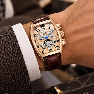 May include: A tonneau-shaped wristwatch with a rose gold-tone case, a champagne-colored dial, and large black Arabic numerals. It features blue hands, a date window at the 12 o'clock position, two sub-dials, an open-heart balance wheel, and a brown leather strap.