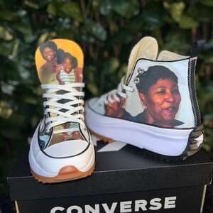 May include: A pair of white high-top Converse sneakers with a black trim. The sneakers are personalized with photos of people on the sides. The sneakers are sitting on a black Converse shoebox.