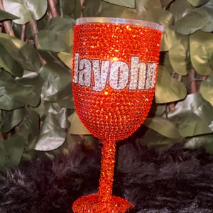 May include: A red wine glass covered in orange rhinestones with the name "Jayohna" spelled out in white rhinestones.