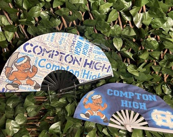 May include: Two decorative folding fans with the text "Compton High" in blue and white. One fan has a repeating pattern of the text, and the other has a cartoon mascot. The fans are set against a backdrop of green leaves.